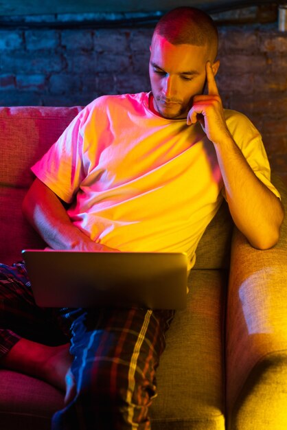 Portrait of handsome young Caucasian man sitting at home in neon orange pink lighted room.