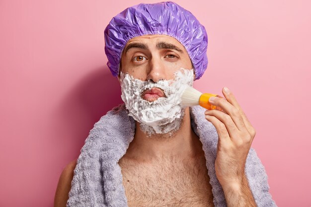 Portrait of handsome young European man applies shaving foam on face with brush, prepares for shave, wears showercap, has soft towel around neck, stands topless indoor. Masculine skin care concept