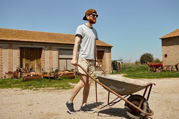 Portrait of handsome young male hipster in snapback and sunglasses carrying trolley from warehouse