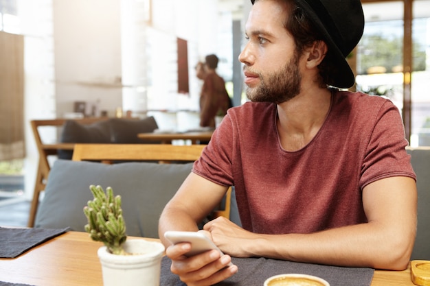 Портрет красивого молодого мужчины с щетиной, сидящего за деревянным столом и держащего универсальный смартфон