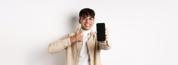 Free photo portrait of handsome young man in glasses showing empty smartphone screen and thumb up recommending