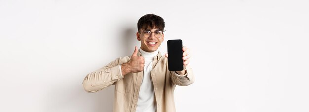 Portrait of handsome young man in glasses showing empty smartphone screen and thumb up recommending