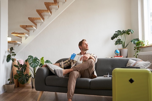 Free photo portrait of handsome young man sits with suitcase in living room holds two plane tickets going on