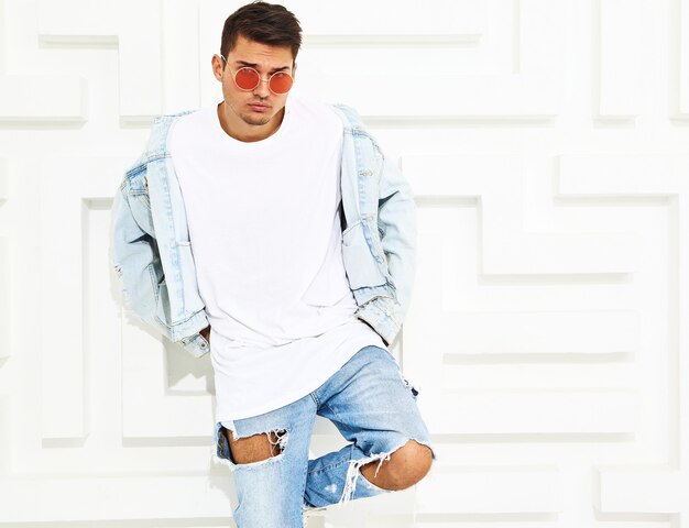 Portrait of handsome young model man dressed in jeans clothes posing near white textured wall