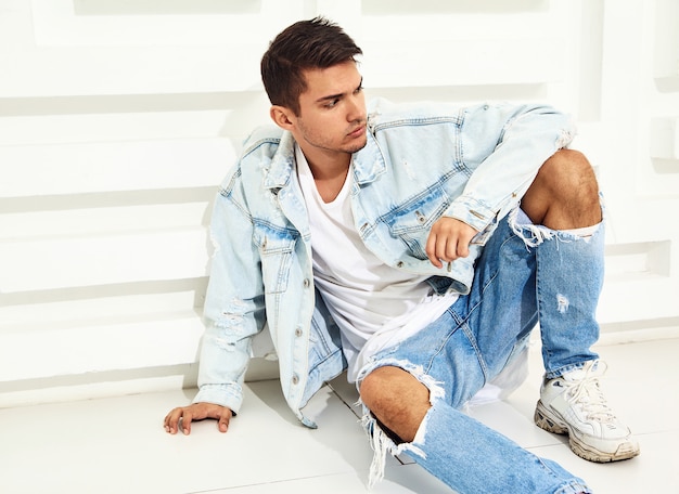 Portrait of handsome young model man dressed in jeans clothes sitting near white textured wall