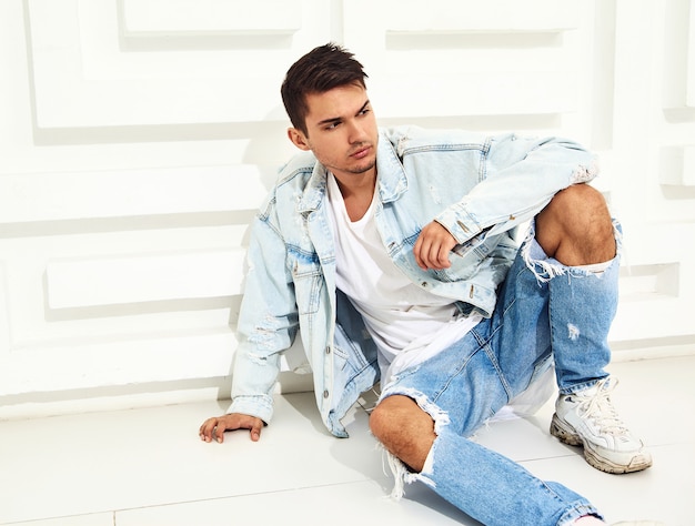 Портрет красивого молодого модельного человека, одетого в джинсовую одежду, сидит возле белой фактурной стены