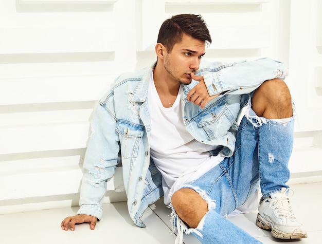 Portrait of handsome young model man dressed in jeans clothes sitting near white textured wall