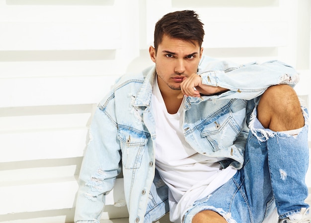 Portrait of handsome young model man dressed in jeans clothes sitting near white textured wall