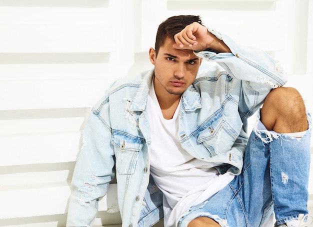 Portrait of handsome young model man dressed in jeans clothes sitting near white textured wall