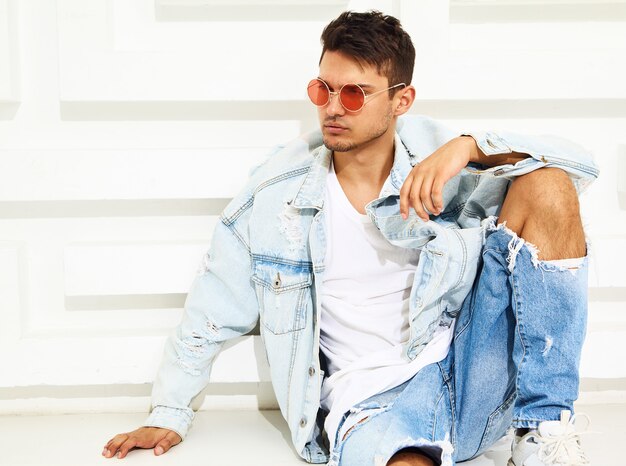 Portrait of handsome young model man dressed in jeans clothes sitting near white textured wall