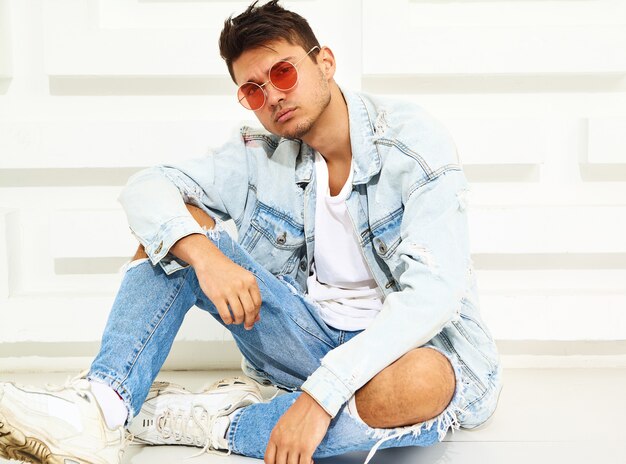 Portrait of handsome young model man dressed in jeans clothes sitting near white textured wall