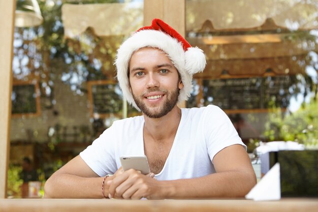 Portrait of handsome young unshaven man wearing red hat with white fur holding mobile phone