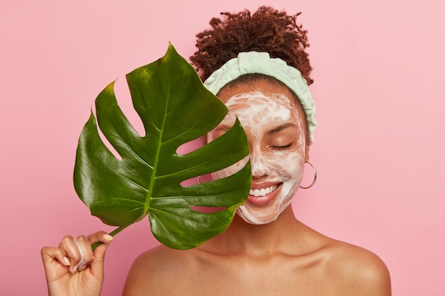 Portrait of happy African American woman covers half of face with green leaf, cleans face, washes with bubble soap, stands topless, cares aboout her beauty and body
