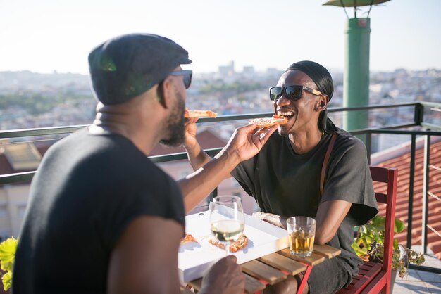 Portrait of happy African gay couple eating pizza. Two happy bearded men sitting at table on faraway city background feeding each other slices of pizza. Same sex love and LGBT couples rights concept