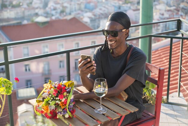 옥상에서 데이트를 기다리는 행복한 아프리카 남자의 초상화. 티셔츠와 선글라스를 쓴 웃고 있는 남자가 꽃과 전화기를 들고 발코니에 있는 탁자에 앉아 그것을 바라보고 있습니다. 진정한 사랑과 데이트 개념