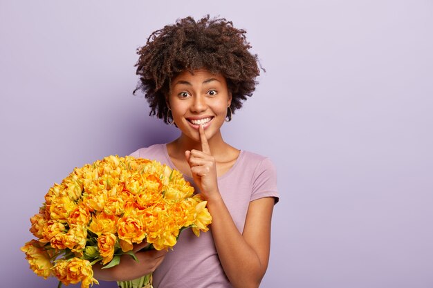 Portrait of happy afro american woman makes silence gesture with delighted expression, holds big bunch of spring flowers