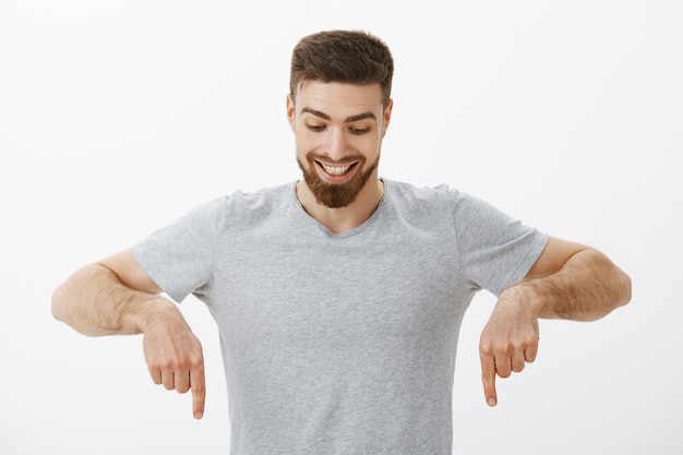 Free photo portrait of happy and amazed good-looking masculine male with perfect hairstyle and beard looking and pointing down with delight and curiosity enjoying watching downwards at interesting copy space