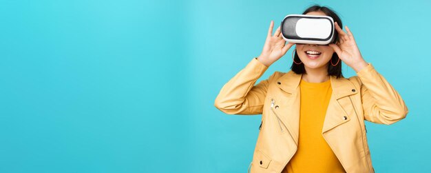 Portrait of happy asian female model using VR headset smiling and laughing in virtual reality glasses standing over blue background