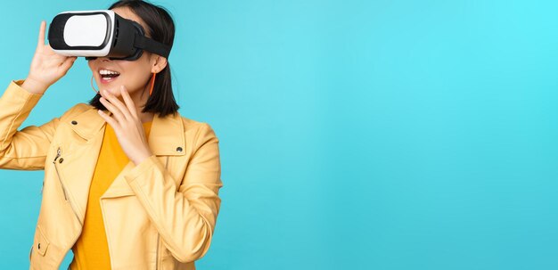 Portrait of happy asian female model using VR headset smiling and laughing in virtual reality glasses standing over blue background