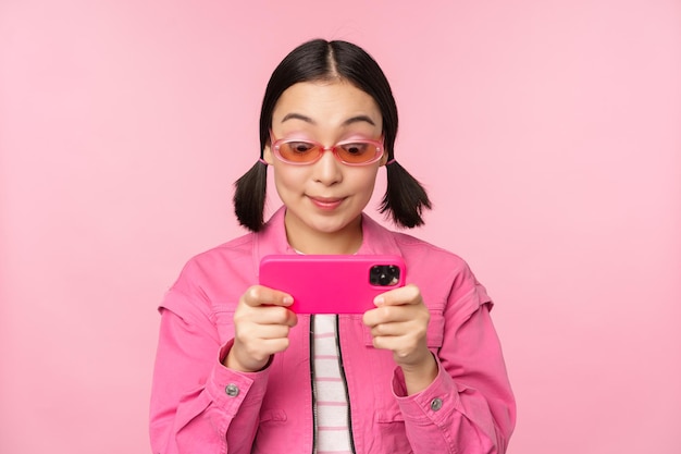 Portrait of happy asian girl playing on smartphone watching videos on mobile phone app standing over pink background