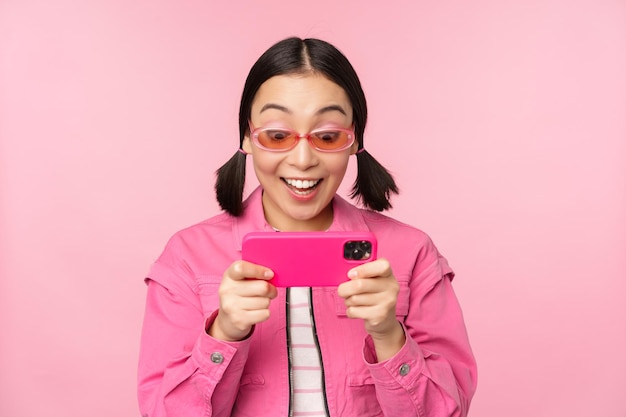 분홍색 배경 위에 서 있는 휴대전화 앱에서 동영상을 보고 스마트폰으로 노는 행복한 아시아 소녀의 초상화