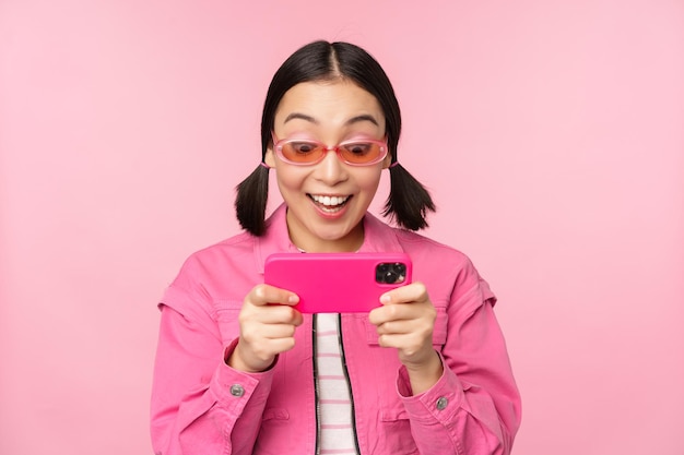 Portrait of happy asian girl playing on smartphone watching videos on mobile phone app standing over pink background