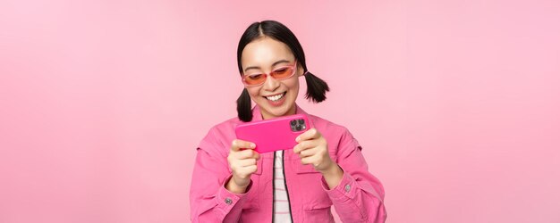 Portrait of happy asian girl playing on smartphone watching videos on mobile phone app standing over pink background