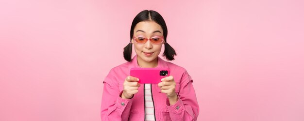 立っている携帯電話アプリでビデオを見てスマートフォンで遊んで幸せなアジアの女の子の肖像画