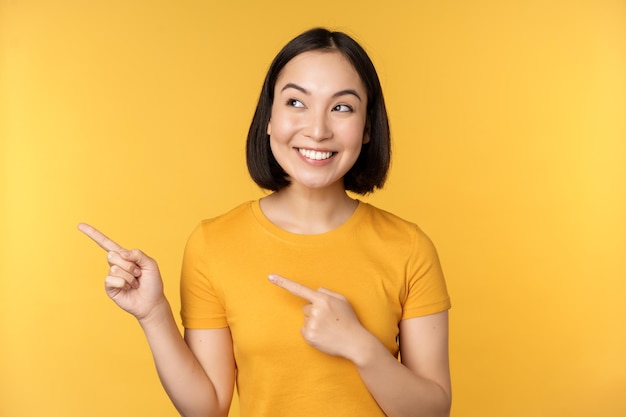 Портрет счастливой азиатской девушки, указывающей пальцами и смотрящей влево, улыбающейся, изумленно проверяющей промо-баннер с рекламой на желтом фоне