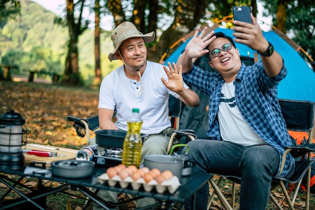 Portrait happy Asian man friends Making a video call with smartphone in camping Cooking set front ground Outdoor cooking traveling camping lifestyle concept