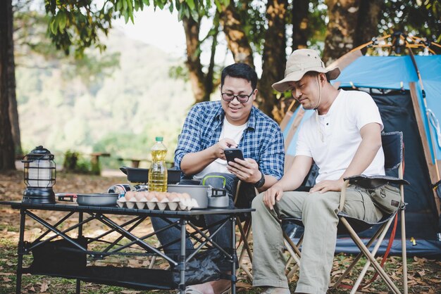 Portrait happy Asian man friends sitting on chair in the camp with talking and looking at smartphone Cooking set front ground Outdoor cooking traveling camping lifestyle concept