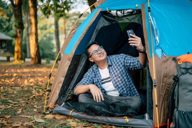 Portrait happy Asian man glasses making a video call with smartphone in tent camping Cooking set front ground Outdoor cooking traveling camping lifestyle concept