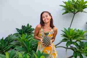 Free photo portrait of happy asian smiling woman in yellow summer dress hold pineapple