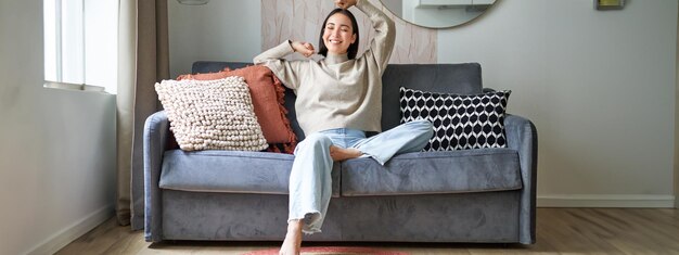 Portrait of happy asian woman feeling lazy stretching on sofa and smiling pleased relaxing at home
