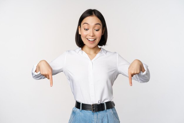 Portrait of happy asian woman pointing fingers down and looking below at advertisement showing info banner advertising standing over white background