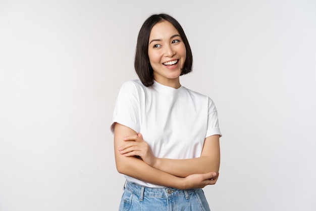 Портрет счастливой азиатки, улыбающейся, позирующей уверенно скрещенными руками на груди, стоящей на фоне студии