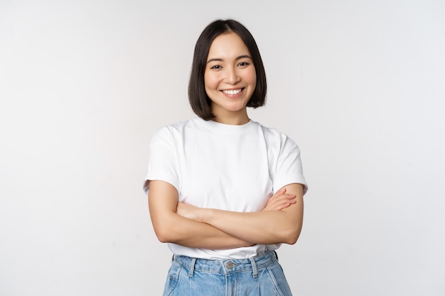 Портрет счастливой азиатки, улыбающейся, позирующей уверенно скрещенными руками на груди, стоящей на фоне студии