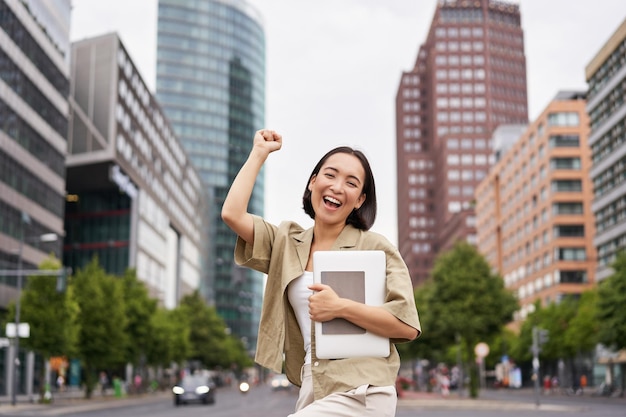 幸せなアジアの女性の肖像画は、通りの道路の近くにタブレットを持って立ち、手を上げて応援しています