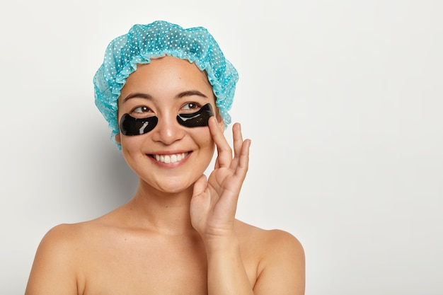Portrait of happy Asian woman with dark patches for skin care under eyes, has recovery treatment on face, wears blue showercap, stands bare over white wall, removes wrinkles and puffiness