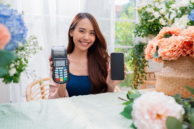 Portrait of a happy asian young adult florist floral shop with cheerful smiling hand show smartphone small business ownerFlorist showing a smartphone while surrounded with flowers and plants