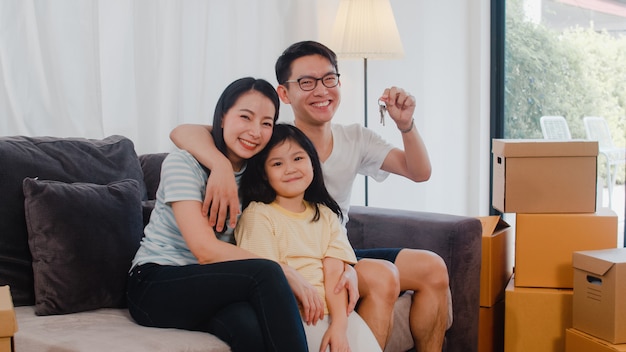 Portrait of Happy Asian young family bought new house. Japanese little preschool daughter with parents mother and father holds in hand keys sitting on sofa in living room smiling looking at camera.