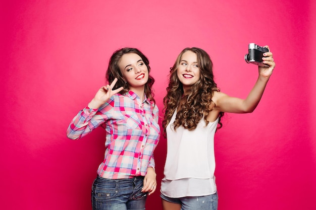 Portrait of happy attractive girlfriends with long wavy hairstyles taking selfportrait via old film camera against red background Brunette in plaid shirt making bunny ears to her friend