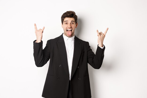 Портрет счастливого привлекательного парня, весело проводящего время на вечеринке, в черном костюме, показывающего знак рок-н-ролла и язык, возбужденного на белом фоне