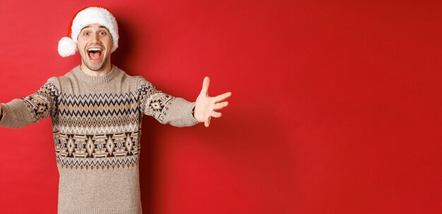 Portrait of happy, attractive man in christmas sweater and santa hat, reaching hands for gift, want to take something and smiling, standing over red background