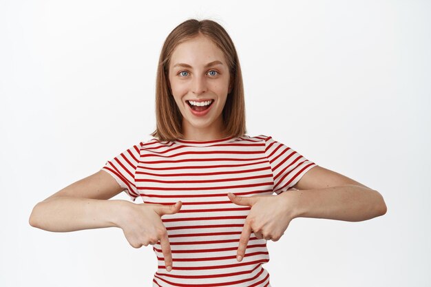 Portrait of happy attractive young woman pointing fingers down, showing link or advertisement banner, sale info, recommend follow page, standing over white background. Copy space