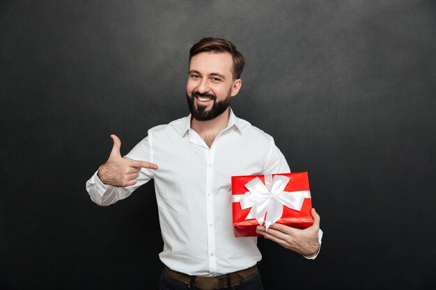 Portrait of happy bearded man holding red gift box and pointing index finger on it over dark gray wall