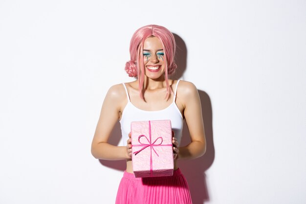 Портрет счастливой красивой девушки-дня рождения, выглядящей взволнованной, получающей подарок на день рождения и радостно улыбающейся, стоящей в розовом парике и праздничном наряде.