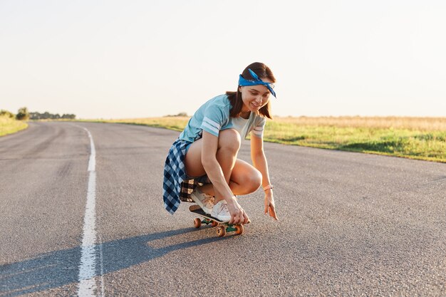 Portrait of happy beautiful dark haired female wearing casual attire and hair band surfing sitting on skateboard, having fun outdoor alone, healthy active lifestyle.