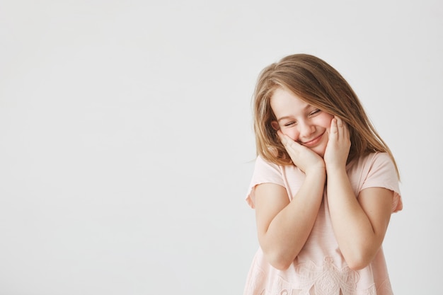 Portrait of happy beautiful girl with blond hair in pink t-shirt squeezing face with hands, closing eyes, feeling shy after cute boy in school made her compliment