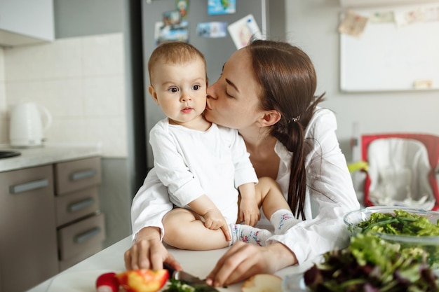 식탁에 앉아 놀란 표정으로 아기가 식탁에 앉아 있는 사랑스러운 아기에게 키스하는 행복한 아름다운 어머니의 초상